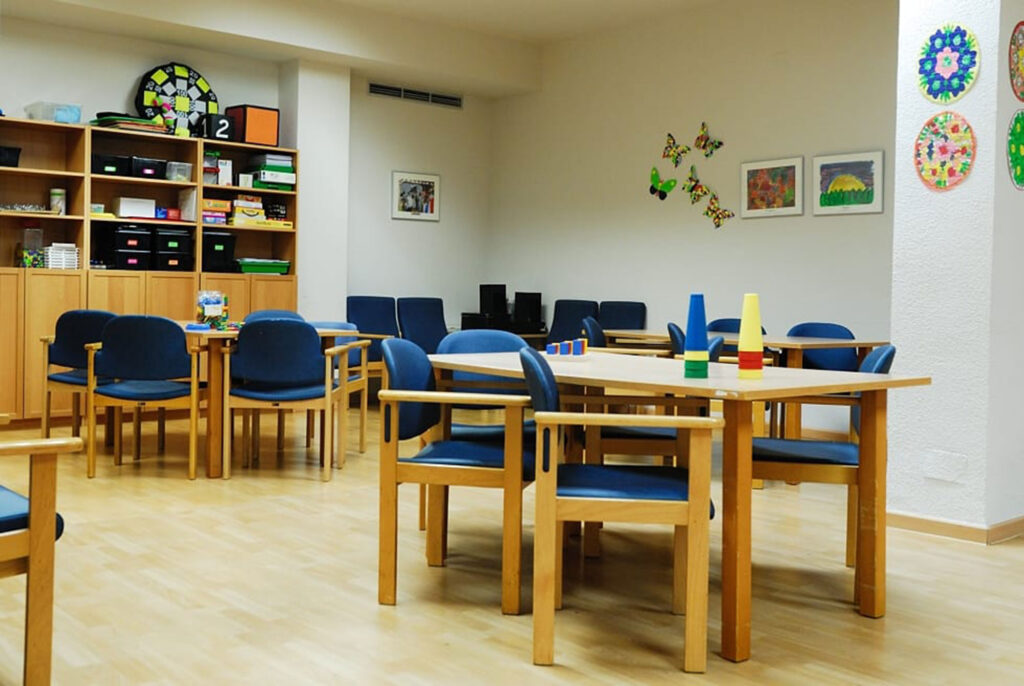 Sala de actividades con varias mesas y sillas, juegos sobre las mesas y estantería con material al fondo