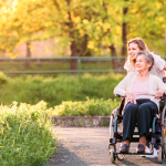 Persona mayor en silla de ruedas acompañada de un cuidador paseando por una senda